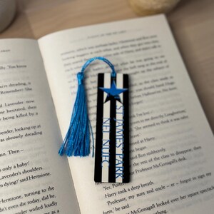 K&ouml;nnte beinhalten: Schwarz-wei&szlig; gestreifter Lesezeichen mit blauem Stern und Quaste. Der Text "ST JAMES PARK" ist auf dem Lesezeichen zu sehen. Das Lesezeichen liegt in einem offenen Buch.