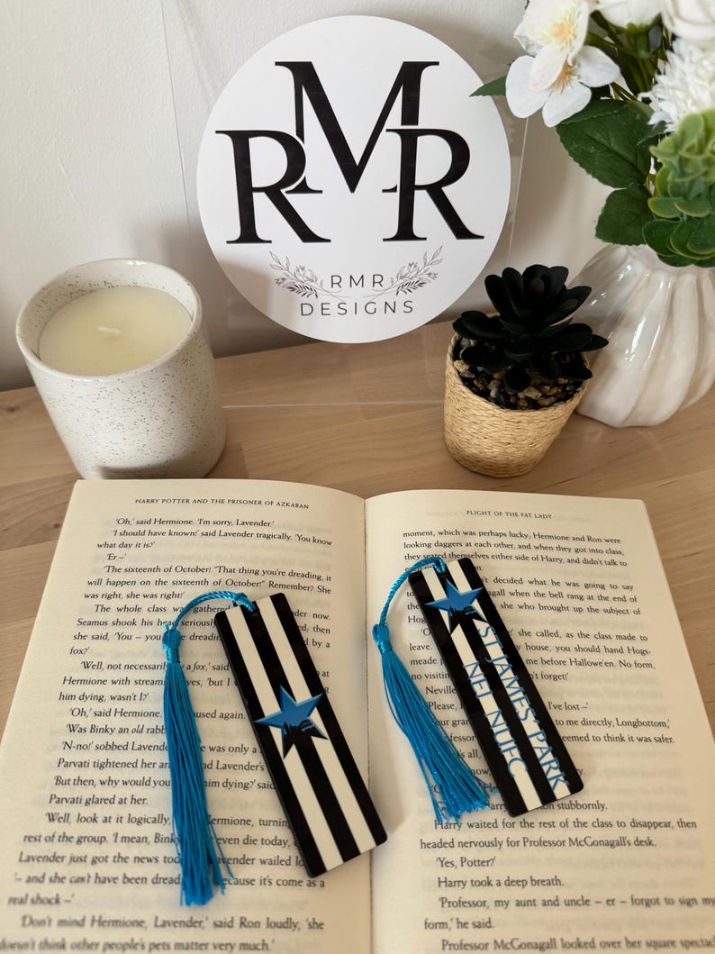 K&ouml;nnte beinhalten: Zwei schwarz-wei&szlig; gestreifte Lesezeichen mit blauen Quasten und einem blauen Sterndesign werden auf einem offenen Buch gezeigt. Die Lesezeichen tragen die Texte "NEL NUFC" und "ST JAMES PARK". Eine Kerze, eine kleine Sukkulente und ein Schild mit dem Text "RMR DESIGNS" befinden sich im Hintergrund.