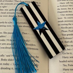 K&ouml;nnte beinhalten: Schwarz-wei&szlig; gestreifter Lesezeichen mit blauem Sterndesign und blauer Quaste. Das Lesezeichen ist rechteckig und zeigt einen Stern mit einer Silhouette im Inneren. Ein dekoratives Accessoire f&uuml;r B&uuml;cherliebhaber.