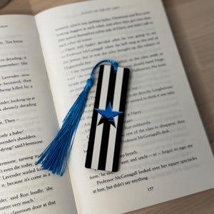 K&ouml;nnte beinhalten: Ein schwarz-wei&szlig; gestreifter Lesezeichen mit blauem Stern und Quaste, das auf einem offenen Buch liegt. Der Lesezeichen ist rechteckig und hat vertikale Streifen. Die Quaste ist leuchtend blau.