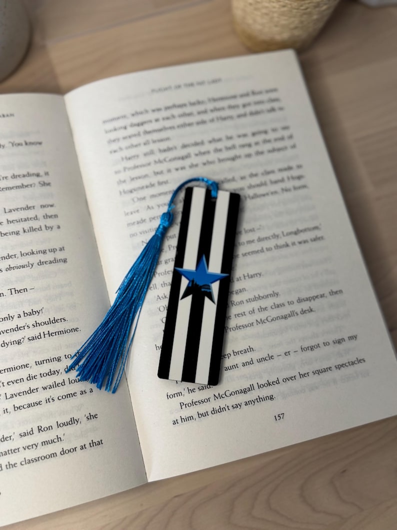 K&ouml;nnte beinhalten: Ein schwarz-wei&szlig; gestreifter Lesezeichen mit blauem Stern und Quaste, das auf einem offenen Buch liegt. Der Lesezeichen ist rechteckig und hat vertikale Streifen. Die Quaste ist leuchtend blau.