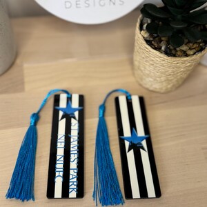 K&ouml;nnte beinhalten: Zwei schwarz-wei&szlig; gestreifte Lesezeichen mit blauen Quasten. Jedes Lesezeichen hat einen blauen Stern und den Text "ST JAMES PARK NUFC" in Blau. Die Lesezeichen werden auf einer hellen Holzoberfl&auml;che pr&auml;sentiert.