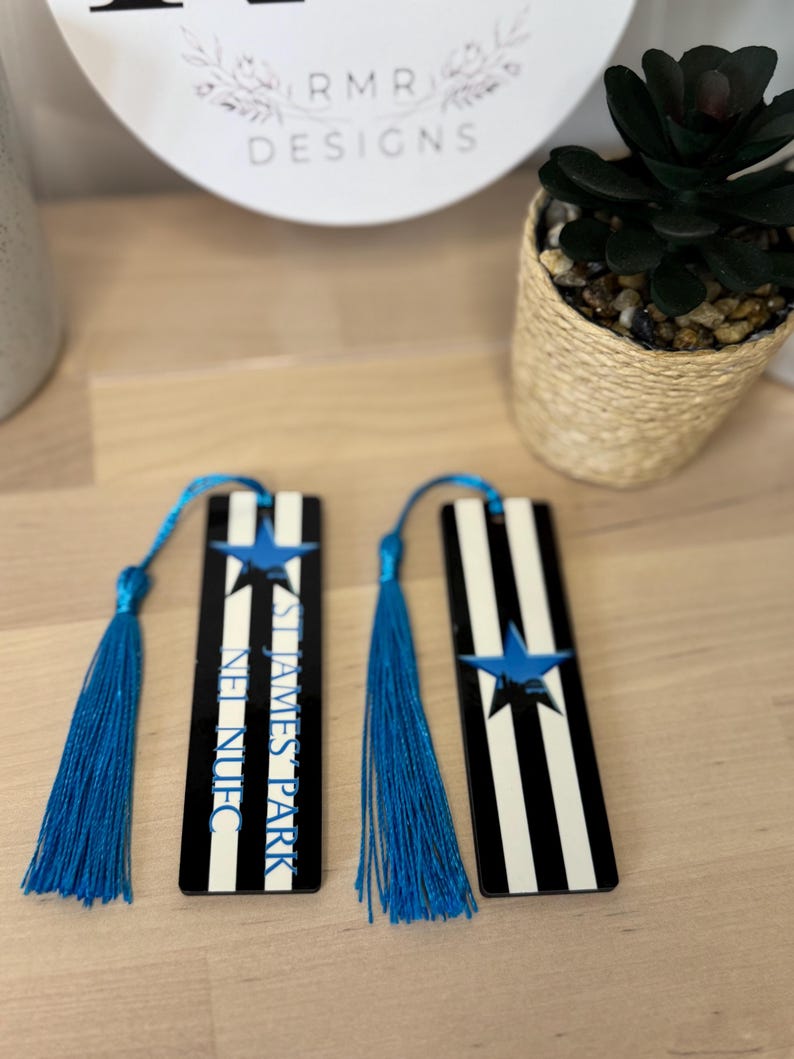 K&ouml;nnte beinhalten: Zwei schwarz-wei&szlig; gestreifte Lesezeichen mit blauen Quasten. Jedes Lesezeichen hat einen blauen Stern und den Text "ST JAMES PARK NUFC" in Blau. Die Lesezeichen werden auf einer hellen Holzoberfl&auml;che pr&auml;sentiert.