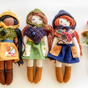 May include: Three handmade fabric dolls with different colored hair, clothing, and hats. The dolls are wearing skirts with pockets and have crocheted scarves. The dolls are standing on a white background.