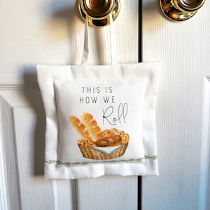 May include: A white fabric door hanger with the text "THIS IS HOW WE Roll" and a watercolor illustration of bread in a basket. The hanger is suspended from a gold doorknob.