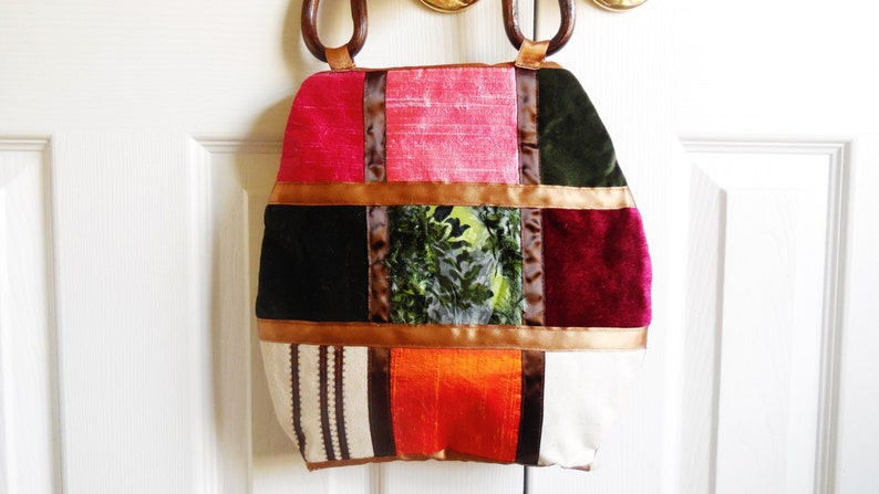 May include: A patchwork tote bag with a variety of colors and textures, including pink, red, green, brown, and white. The bag has a unique design with different fabrics sewn together in a square pattern. The bag has wooden handles.