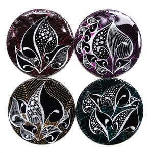 Puede incluir: Cuatro posavasos redondos con fondo negro y diseños florales blancos. Cada posavasos tiene un fondo de color diferente: rojo, morado, dorado y verde.