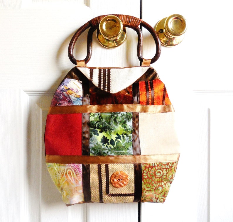 May include: A patchwork tote bag with a brown wooden handle. The bag is made of different fabrics in shades of brown, orange, green, and white. The bag has a large button on the front.