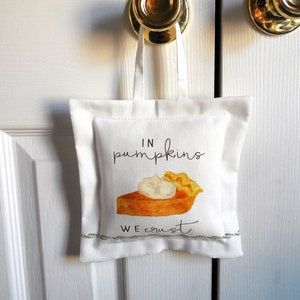 May include: White fabric bag with a handle, featuring a watercolor illustration of pumpkin pie with whipped cream. The text "IN pumpkins WE crust" is written in a script font. The bag is hanging on a white door.