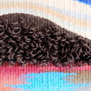 May include: Close-up of a woven tapestry with a dark brown, curly fringe. The tapestry features a colourful abstract design with shades of blue, pink, and white.