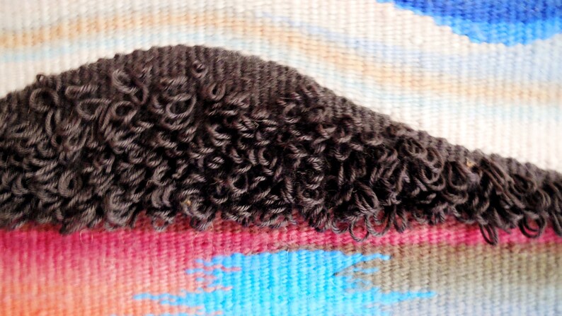 May include: Close-up of a woven tapestry with a dark brown, curly fringe. The tapestry features a colourful abstract design with shades of blue, pink, and white.