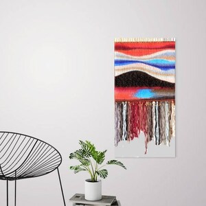 May include: A colourful woven wall hanging with a red, blue and brown colour scheme. The hanging has a fringe border and a textured surface.
