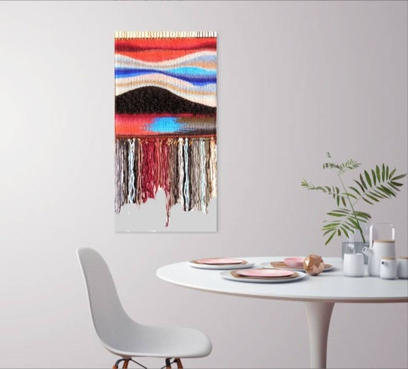 May include: A colourful woven wall hanging with a geometric pattern and fringe. The hanging is made of various colours of yarn, including red, blue, white, and black.