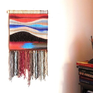 May include: A colourful woven wall hanging with a geometric design featuring blue, red and brown hues. The hanging has a fringe border and is made with a variety of yarns.