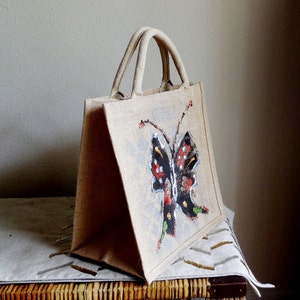 Puede incluir: Una bolsa de mano beige con un diseño de mariposa pintado. La mariposa es negra con manchas rojas y blancas y detalles verdes.