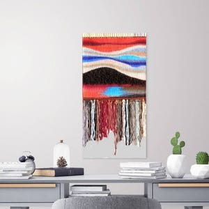 May include: A colourful woven wall hanging with a red, blue and white pattern. The hanging has a fringe of yarn at the bottom.