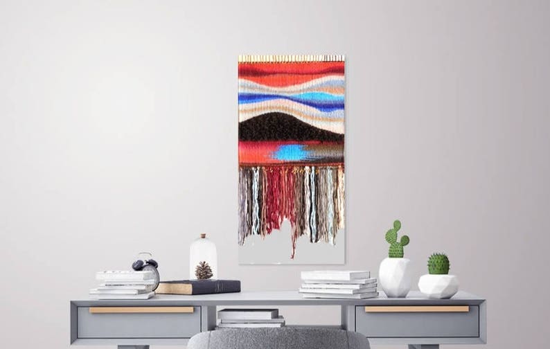 May include: A colourful woven wall hanging with a red, blue and white pattern. The hanging has a fringe of yarn at the bottom.