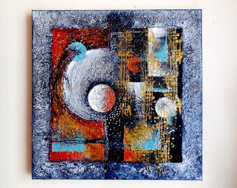 May include: Abstract square painting with a textured, dark blue border. The artwork features a central white circle with red and silver accents, and a mix of red, orange, gold, and blue geometric shapes. The painting is mounted on a white wall.
