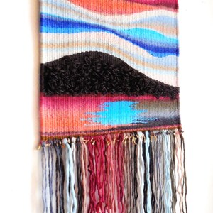 May include: A woven wall hanging with a colourful abstract design featuring blue, red and brown hues. The hanging has a fringe of yarn in various colours.