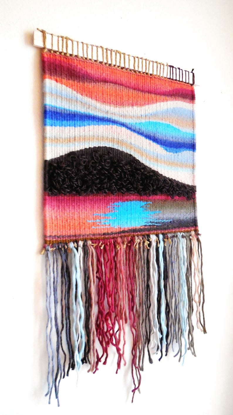 May include: A woven wall hanging with a colourful abstract design featuring blue, red and brown hues. The hanging has a fringe of yarn in various colours.