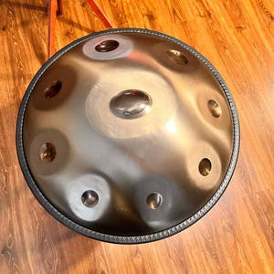 May include: A steel handpan drum with a bronze finish. The instrument has a circular shape with multiple dimples and a central dome. A black rim surrounds the edge of the drum. The handpan sits on a wooden floor.