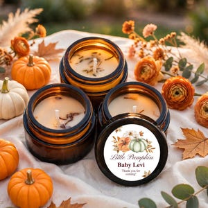 May include: Three amber glass jars containing cream-colored wax candles with visible wicks. One lid features a circular label with a watercolor pumpkin illustration and the text "Little Pumpkin Baby Levi Thank you for coming!" surrounded by autumn decor.