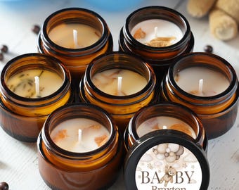 May include: A collection of six amber glass jars containing white wax candles with cotton wicks. One lid features a circular label with a teddy bear illustration, the text BABY Braxton, and the date 06.09.24, with the phrase Thank you for coming.