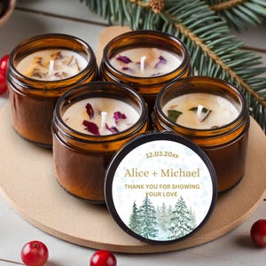 May include: Four amber glass jar candles with white wax and botanical inclusions. One black lid features a circular label with the text Alice + Michael, 12.03.20xx, and THANK YOU FOR SHOWING YOUR LOVE above a printed illustration of green pine trees.