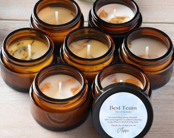 May include: Eight small amber glass jars with lids, filled with white candles. The jars are arranged in a circle on a light wood surface. One jar has a black lid with the text "Best Team" and a short message about teamwork.