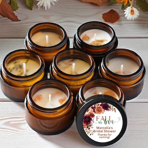 Puede incluir: Un conjunto de siete frascos pequeños de vidrio ámbar rellenos de velas de cera blanca, cada una con una mecha centrada. Una tapa está colocada en primer plano, mostrando un gráfico floral con el texto Fall in love, Marcella's Bridal Shower, Thanks for coming.
