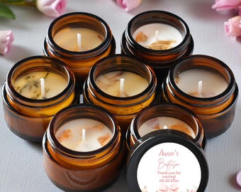 May include: Six small amber glass jars containing white wax candles with visible wicks. One black lid is placed in front, featuring a circular white label with pink text reading Annie's Baptism, Thank you for coming, 04.20.2024, and floral art.