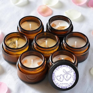 May include: A collection of six small amber glass jars containing white wax candles with visible wicks. One black lid is placed in front, featuring a circular label with two heart outlines, the letters L and C, and the date 8.20.20XX.