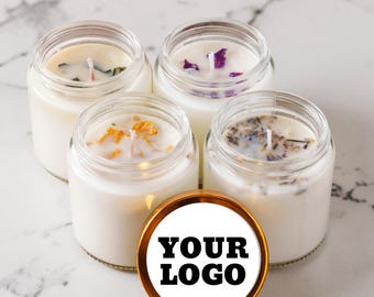 Könnte beinhalten: Vier durchsichtige Gläser gefüllt mit weißen Wachskerzen, jede mit einem Docht und getrockneten botanischen Elementen versehen. Ein runder, goldgeränderter Deckel mit dem schwarzen Aufdruck YOUR LOGO liegt vor den Kerzen.