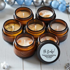 May include: A collection of small, amber-colored glass jar candles with white wax and wicks. One candle has a black lid with the text "Oh Baby!" and a personalized message. The candles are arranged on a white wooden surface.