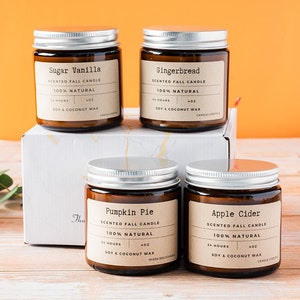May include: Four scented fall candles in glass jars with silver lids, presented in a white box. The labels read "Sugar Vanilla", "Gingerbread", "Pumpkin Pie", and "Apple Cider". The background is orange and the candles are on a wooden surface.