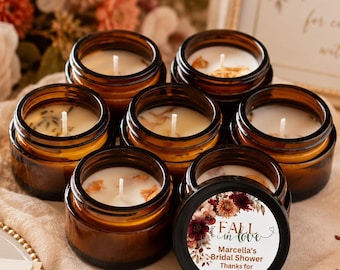 May include: A collection of seven amber glass jar candles with white wax and cotton wicks. One candle features a circular label with floral graphics and the text: Fall in love, Marcella's Bridal Shower, Thanks for coming. The candles are arranged together.