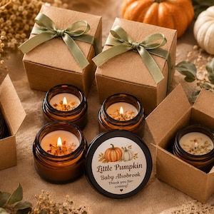 Può includere: Diversi barattoli in vetro ambrato contenenti candele bianche con stoppini accesi, alcuni inseriti in scatole di cartone marrone aperte con nastri verde salvia. Un coperchio nero presenta un'illustrazione di zucca e il testo Little Pumpkin Baby Alsobrook Thank you for coming.