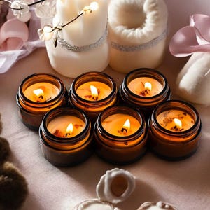 May include: A collection of lit candles in small, brown glass jars. The candles are arranged on a light pink surface, surrounded by decorative items like a plush bear, a wrapped gift, and other candles.