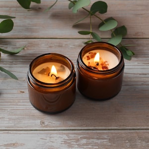 May include: Two amber glass candle jars with a natural wax candle inside. The candles are lit and the flames are visible. The jars are on a light brown wooden surface.