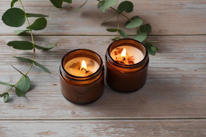 May include: Two amber glass candle jars with a natural wax candle inside. The candles are lit and the flames are visible. The jars are on a light brown wooden surface.