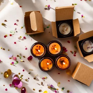 May include: Overhead shot of several small, round candles in black containers, some lit with orange flames. The candles are in and out of small, open cardboard boxes. The scene is decorated with colorful confetti.