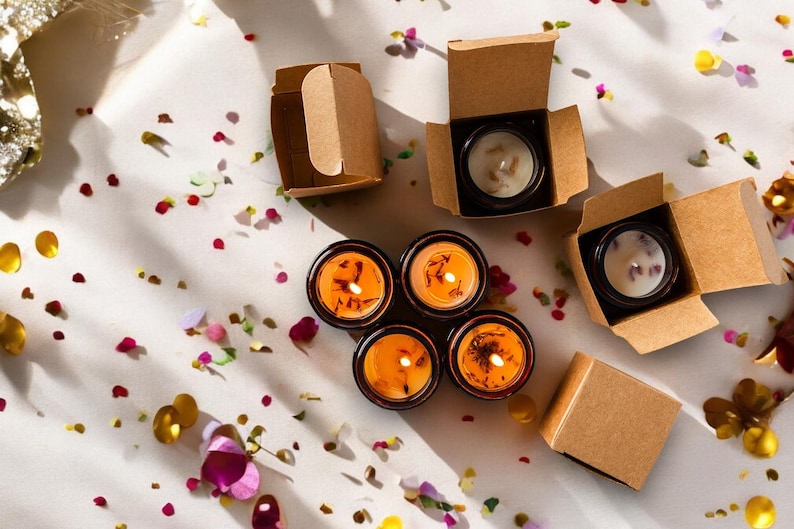 May include: Overhead shot of several small, round candles in black containers, some lit with orange flames. The candles are in and out of small, open cardboard boxes. The scene is decorated with colorful confetti.