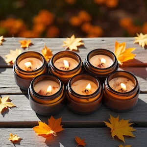 May include: Five amber glass jars containing lit candles with visible wicks and wax, arranged on a wooden surface alongside several small orange pumpkins and scattered yellow and orange maple leaves, creating a warm and seasonal display.