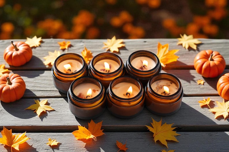 May include: Five amber glass jars containing lit candles with visible wicks and wax, arranged on a wooden surface alongside several small orange pumpkins and scattered yellow and orange maple leaves, creating a warm and seasonal display.