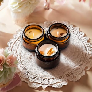 May include: Three small, amber-colored glass jar candles with white wax and dried botanicals sit on a white lace doily. The candles are surrounded by cupcakes and flowers, creating a warm and inviting scene.