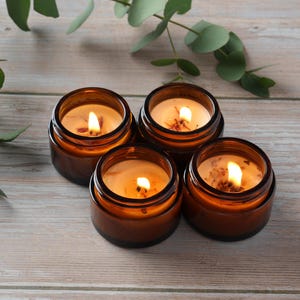 May include: Four amber glass candle jars with lids, each containing a lit candle with a warm, yellow glow. The candles are arranged in a circle on a light wood surface.
