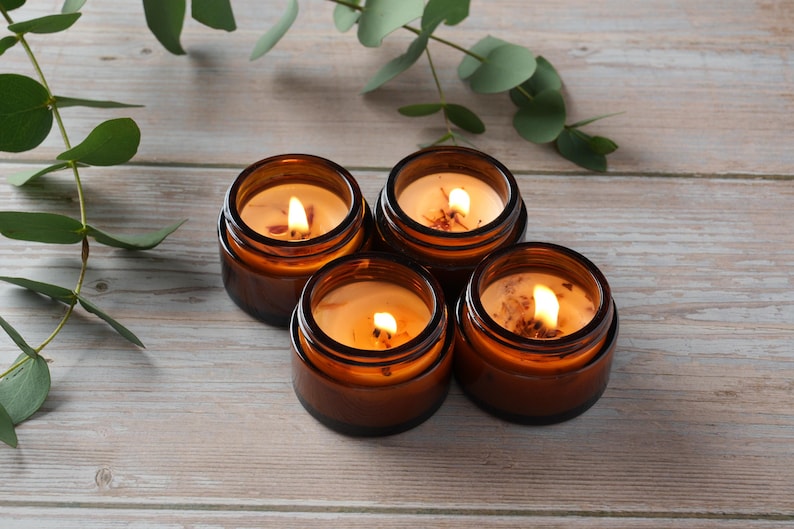 May include: Four amber glass candle jars with lids, each containing a lit candle with a warm, yellow glow. The candles are arranged in a circle on a light wood surface.