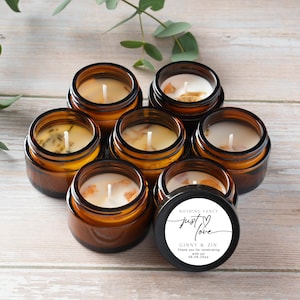 May include: Seven small, round amber glass jars filled with wax candles. The candles have white wicks and are surrounded by decorative elements. One jar has a black lid with the text "Nothing Fancy just love Ginny & Zin".