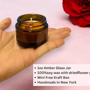 May include: A small amber glass jar candle with a white wick, held in a hand. The candle is filled with soy wax and dried flower petals. A red rose is in the background.