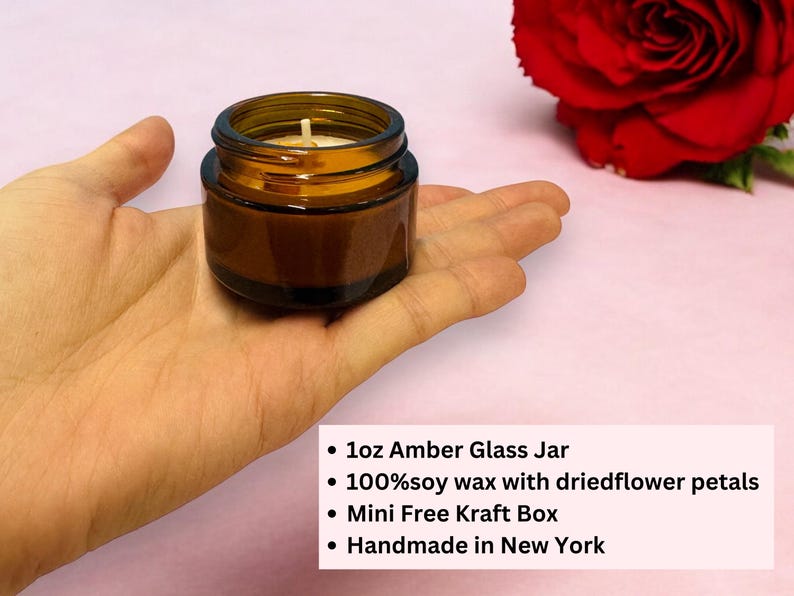 May include: A small amber glass jar candle with a white wick, held in a hand. The candle is filled with soy wax and dried flower petals. A red rose is in the background.